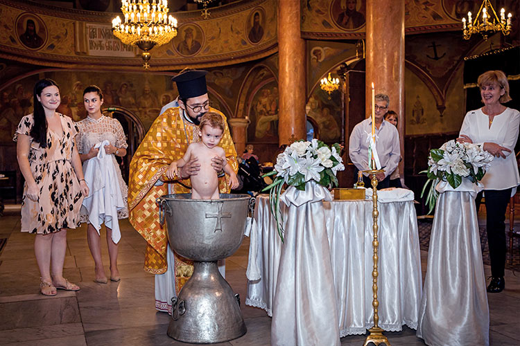 Отецът кръщава момченце в купела по време на ритуал Свето Кръщение в катедрален храм Света Богородица, град Пловдив.