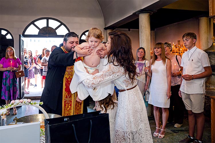 Кръстницата приема момиченцето след кръщаването в купела от отеца по време на ритуал Свето Кръщение в храм Света Троица, Кръстова Гора.