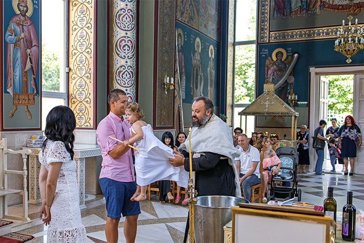 Кръстникът приема момиченцето след кръщаването в купела от отеца по време на ритуал Свето Кръщение в храм Свети Николай Мирликийски Чудотворец, град Пловдив.
