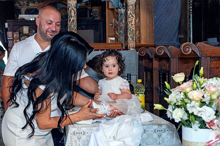 Кръстницата обува новопокръстено момиченце облечено в новата си бяла рокличка след кръщаване в купела по време на ритуал Свето Кръщение в храм Свети Свети Константин и Елена. град Пловдив.